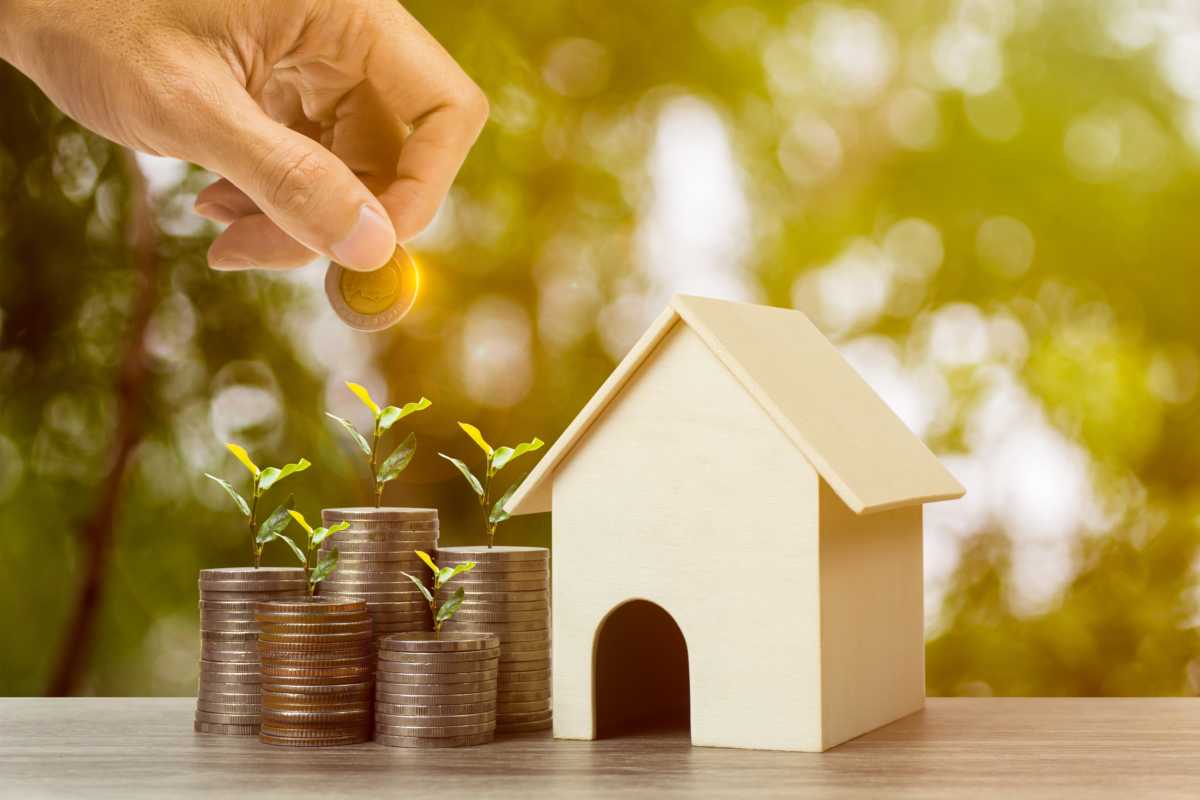 Property investment or saving money for buy home. A businessman hand holding coin over growing plant on stacked coins and resident house model on wooden table.